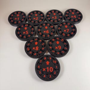 Puede incluir: Una colección de diez fichas de juego circulares negras con números y flechas rojas. Cada ficha tiene un número diferente de x1 a x10, con un diseño similar a un engranaje en los bordes. Las fichas están dispuestas sobre una superficie blanca.