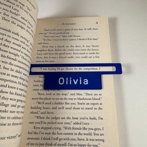 Pode incluir: Um marcador de livros azul com o nome "Olivia" em letras brancas está colocado nas páginas de um livro aberto. O marcador é retangular com uma aba. O livro mostra texto impresso.