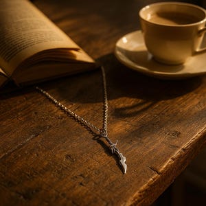 May include: A silver pendant necklace featuring a sword design. The necklace is displayed on a wooden surface, alongside an open book and a cup of coffee. The sword pendant has intricate detailing and a polished finish.