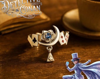 May include: A gold-toned ring featuring a central blue gemstone set within a rose motif, flanked by a crescent moon and playing card suit symbols. A small triangular charm with a clover cutout hangs from the band, which displays Detective Conan Kid text.
