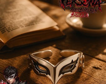 May include: A silver-toned open ring featuring a stylized mask design with black enamel detailing around the eye cutouts. The ring is positioned on a wooden surface near a book and a cup, with the text Persona Amamiya Ren Joker visible.