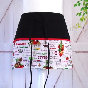 May include: Black and red kitchen apron with a white print of a chili recipe and ingredients. The apron has a tie at the waist and a pocket on the front.