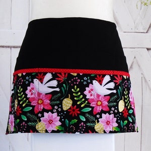 May include: Black apron with a red and white polka dot trim and a festive floral print featuring white doves, red and pink flowers, and green leaves on a black background.