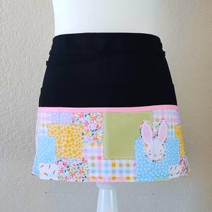May include: Black waist apron with a patchwork design. The patchwork features floral, gingham, and polka dot patterns in pastel colors. A pink bunny is on the front. The apron has a pink trim.