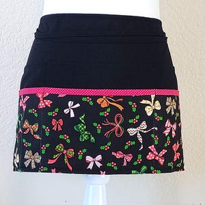 May include: A black half apron with a festive Christmas bow pattern. The apron features a black waistband and a bottom section adorned with colorful bows, holly, and berries on a black background. A pink polka dot trim adds a pop of color.