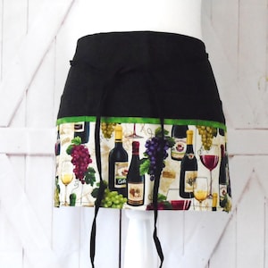 May include: Black apron with a green and white wine bottle print. The apron has a black waistband and ties.