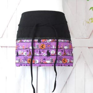 May include: A black and purple apron with a Halloween-themed print. The print features skulls, pumpkins, cats, and other spooky imagery. The apron has a black waistband and ties.