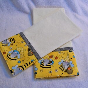 May include: Two yellow fabric tea towels with a white border. The towels feature a pattern of gnomes wearing bee costumes and holding honeycombs. The gnomes are surrounded by bees, flowers, and honeycomb patterns.