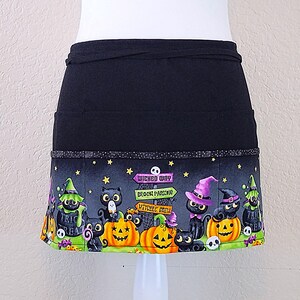 May include: Black Halloween apron with a colorful bottom panel featuring black cats, jack-o'-lanterns, and witch hats. The panel includes text that reads "Wicked Way", "Broom Parking", and "Witches Brew". The apron has two pockets.