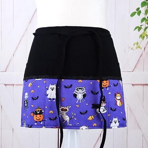May include: A black and purple Halloween-themed apron with a black waistband and a pocket. The apron features a variety of cute cat characters dressed in Halloween costumes, including a ghost, a mummy, a witch, and a skeleton. The apron is perfect for adding a festive touch to any Halloween party or event.