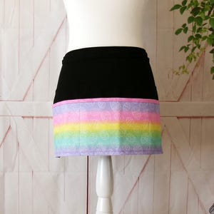 May include: A black apron with a rainbow and white egg pattern on the bottom. The apron has a pink trim.