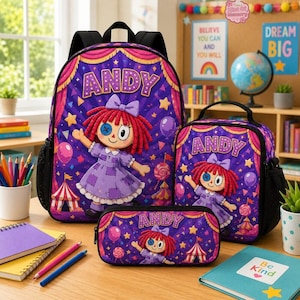 Op de afbeelding: Een paarse rugzak, lunchtas en etui met een cartoonpop met rood haar, een blauw knoop-oog en een paarse jurk. De tekst ANDY staat op elk item gedrukt tegen een achtergrond met een circusthema vol sterren, ballonnen en tenten.
