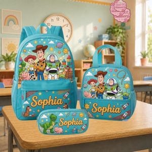 Op de afbeelding: Een lichtblauwe rugzak, een kleinere bijpassende tas en een etui, allemaal voorzien van Toy Story-figuren zoals Woody, Buzz Lightyear en Rex. Elk item is aan de voorzijde gepersonaliseerd met de naam Sophia in een geel cursief lettertype.