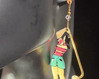 May include: A dangle earring featuring a red heart-shaped stud with a central crystal, connected by a gold-tone chain to a colorful enamel charm of a golfer in a pink striped top and green shorts, swinging a gold-tone golf club with a white golf ball.