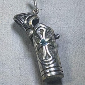 May include: A silver-toned pendant with an ornate design. The pendant features a cylindrical body with a cross-like design and a small, blue gemstone at its center. The top has a decorative, openwork design, and a curved hook for hanging.