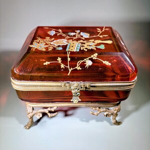 May include: A red-tinted glass jewelry box with a hinged lid, ornate floral designs, and brass accents. The box sits on four decorative brass feet. The lid features gold, blue, and white floral patterns.
