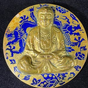 May include: A round, gold-colored medallion featuring a seated Buddha figure. The Buddha is surrounded by blue dragon and floral designs. The medallion has a slightly aged appearance, with some areas showing wear.