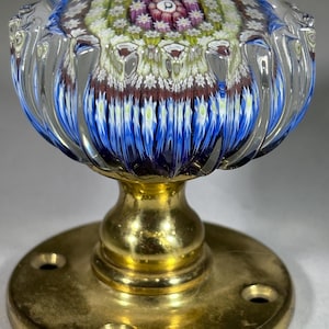 May include: A clear glass doorknob with a colorful floral design. The knob has a brass base and a round brass plate with screw holes. The floral design features blue, white, green, and purple colors.