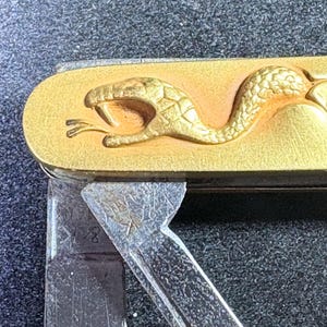 May include: A gold-coloured pocket knife with a snake design. The snake is embossed on the handle. The blade is partially open, showing silver-coloured metal. The knife has a vintage aesthetic.
