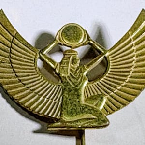 May include: A gold-colored pin featuring an Egyptian goddess with outstretched wings. The goddess is kneeling and holding a sun disc above her head. The wings are detailed with individual feathers.