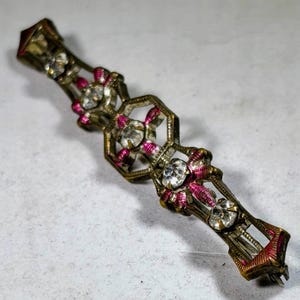May include: Vintage bar pin with a geometric design. The pin features clear rhinestones and pink enamel accents set in a gold-toned metal. The pin is rectangular with decorative elements.