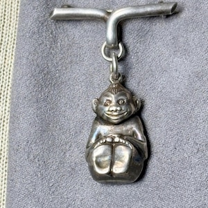 May include: A silver-toned metal brooch featuring a horizontal bar pin at the top. A small, three-dimensional charm shaped like a seated monkey with a smiling face and visible feet hangs from the center of the bar by a circular jump ring.