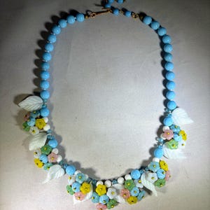 May include: A necklace featuring a strand of light blue spherical beads accented with clusters of small, multi-colored floral charms in yellow, pink, green, and white, interspersed with carved white leaf-shaped beads and a metal clasp.