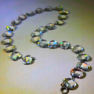 May include: A vintage necklace featuring clear, faceted crystal-like stones. The stones are oval-shaped and linked together with a silver-toned metal chain. The necklace is displayed on a light-colored surface, with the stones reflecting light.
