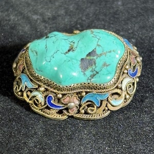 May include: A turquoise stone set in a decorative metal frame. The frame features intricate filigree and colorful enamel accents in blue, pink, and white. The piece has a vintage aesthetic.
