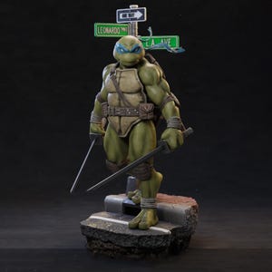 May include: A detailed figurine of Leonardo from Teenage Mutant Ninja Turtles. The figure is green with a blue mask, holding two katanas. It stands on a concrete base with street signs reading "Leonardo" and "LA Ave."