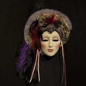 May include: A decorative porcelain mask with a serene expression, adorned with a feathered headdress in shades of purple, black, and burgundy. The mask features a gold and silver halo, pink ribbons, and a jeweled embellishment.