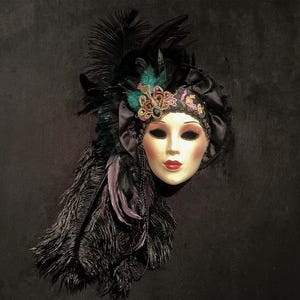 May include: A decorative Venetian mask with a pale face, red lips, and dark eye makeup. The mask is adorned with black feathers, a jeweled headpiece, and a black satin ribbon. A beaded necklace hangs from the mask.