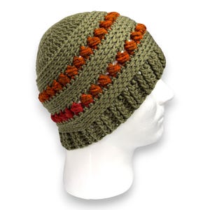 Op de afbeelding: Olijfgroene gebreide beanie met rijen oranje en rode getextureerde accenten. De hoed wordt getoond op een witte mannequinhoofd. De beanie heeft een geribbelde rand en een getextureerd steekpatroon.
