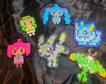 May include: A collection of colorful pixel art figures, including anime-style characters and a moth, crafted from small plastic beads. The figures feature various colors like pink, blue, green, and gray, arranged on a dark fabric background.