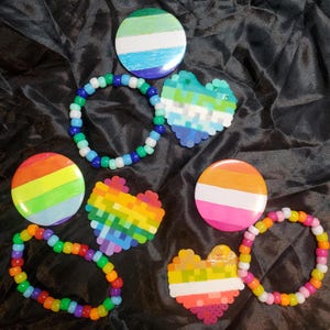 May include: A selection of colourful pride-themed accessories, including round badges and beaded bracelets. The badges feature rainbow stripes and other pride flag designs. The bracelets are made of colourful beads, and there are pixelated heart shapes.