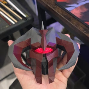 May include: A geometric, dark grey and burgundy object with a red glowing centre, held in a hand. The object has a spiky top and curved horns. The design is angular and appears to be a stylized mask or helmet.