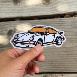 May include: White embroidered patch of a classic sports car with black wheels, a brown windshield, and a black spoiler. The car is outlined in black and has a cream-colored border. The patch is held against a wooden background.