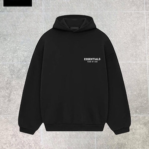 Op de afbeelding: Zwarte hoodie met de tekst "ESSENTIALS FEAR OF GOD" in het wit. De hoodie heeft een capuchon en lange mouwen. Het "DREAM FASHION" logo staat in de linkerbovenhoek.