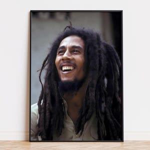 Puede incluir: Retrato enmarcado de un hombre sonriente con largas y oscuras rastas. El retrato está en un marco negro y está colocado contra una pared blanca. El hombre lleva una camisa de color claro.