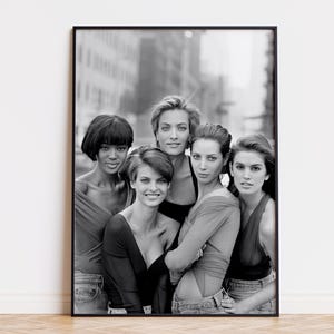 Può includere: Fotografia in bianco e nero incorniciata con cinque donne che posano insieme. Indossano top e jeans. Lo sfondo mostra una scena urbana sfocata. L'immagine è un ritratto.