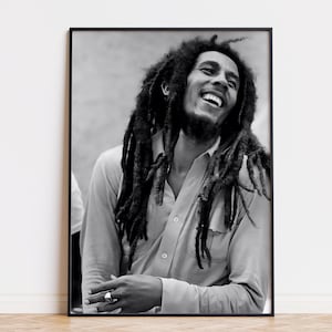 Peut inclure: Portrait encadré en noir et blanc d'un homme souriant avec de longues dreadlocks. Il porte une chemise boutonnée et une bague au doigt. Le portrait est affiché sur un mur blanc au-dessus d'un sol en bois.