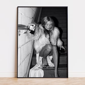 May include: A black and white framed wall art print featuring a person with long hair wearing a textured fur coat and a long skirt, sitting on a staircase. The person holds a small dark object while resting a hand on a metal handrail.