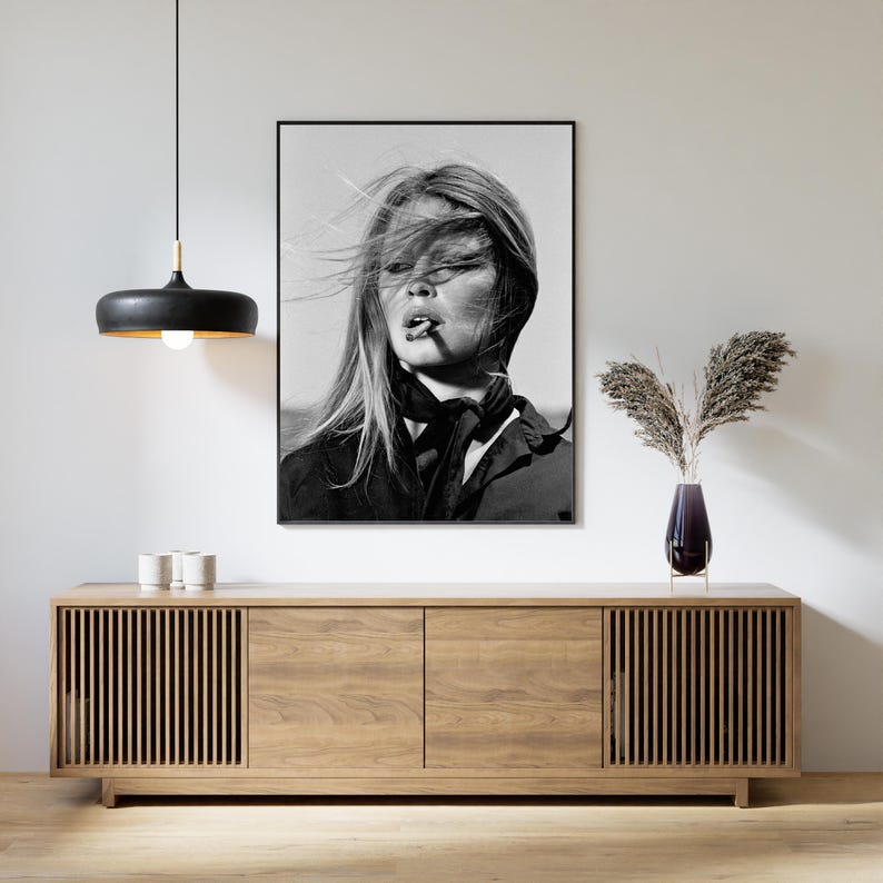 May include: A black and white framed portrait of a person with windblown hair and a dark scarf, hanging above a light brown wooden sideboard with slatted cabinet doors, a black pendant light, and a dark glass vase with dried foliage.