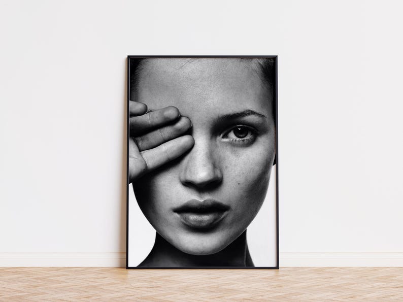 May include: Black and white framed portrait of a person with their hand covering one eye. The image is a close-up, focusing on the face and the hand. The portrait is displayed on a white wall.