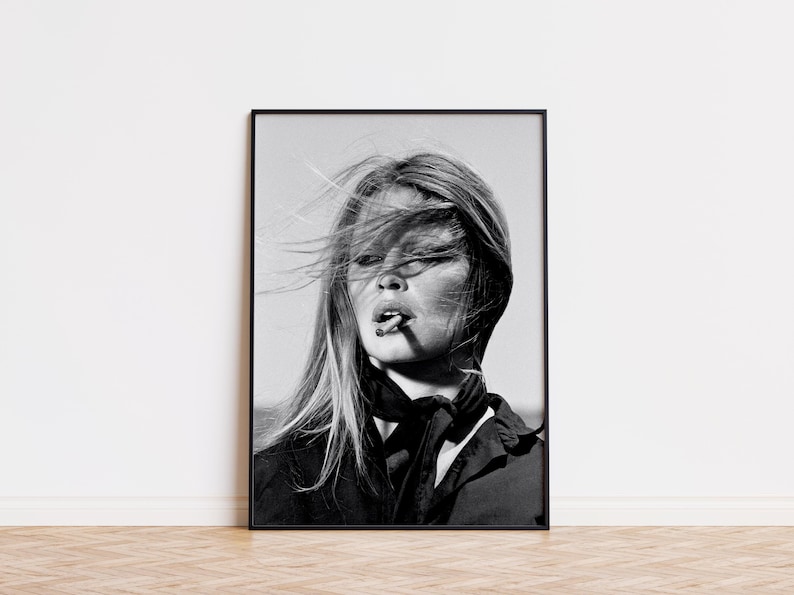 May include: A black and white photographic print in a thin black frame featuring a portrait of a person with long hair blowing in the wind, wearing a dark collared shirt and a tied neck scarf, with a cigarette held between their lips.