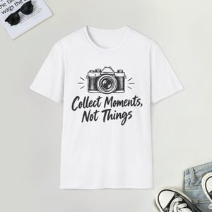 Może przedstawiać: Biały t-shirt z krótkim rękawem z czarną grafiką aparatu vintage umieszczoną na środku klatki piersiowej. Pod aparatem znajduje się napis "Collect Moments, Not Things" wykonany pogrubioną, ciemną i stylizowaną czcionką. Koszulka posiada okrągły dekolt.