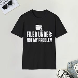 Może przedstawiać: Czarny t-shirt z krótkim rękawem z białą grafiką przedstawiającą ikonę folderu nad napisem "FILED UNDER: NOT MY PROBLEM", wydrukowanym pogrubioną czcionką bezszeryfową na środku klatki piersiowej odzieży.