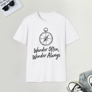 May include: A white short-sleeve t-shirt featuring a black graphic of a compass centered on the chest. Below the compass, the text reads "Wander Often, Wonder Always" in a black, stylized, cursive-inspired font. The shirt has a crew neckline.