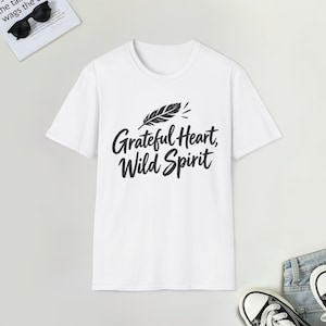 May include: A white short-sleeved t-shirt featuring a black graphic print on the chest. The design includes a stylised feather icon positioned above the text "Grateful Heart, Wild Spirit" written in a bold, cursive, black script font.