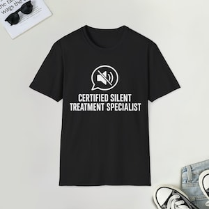 Peut inclure: Un t-shirt noir à manches courtes avec un motif blanc représentant une bulle de dialogue contenant une icône de haut-parleur barré. Sous l'icône, le texte "CERTIFIED SILENT TREATMENT SPECIALIST" est écrit en lettres capitales blanches.